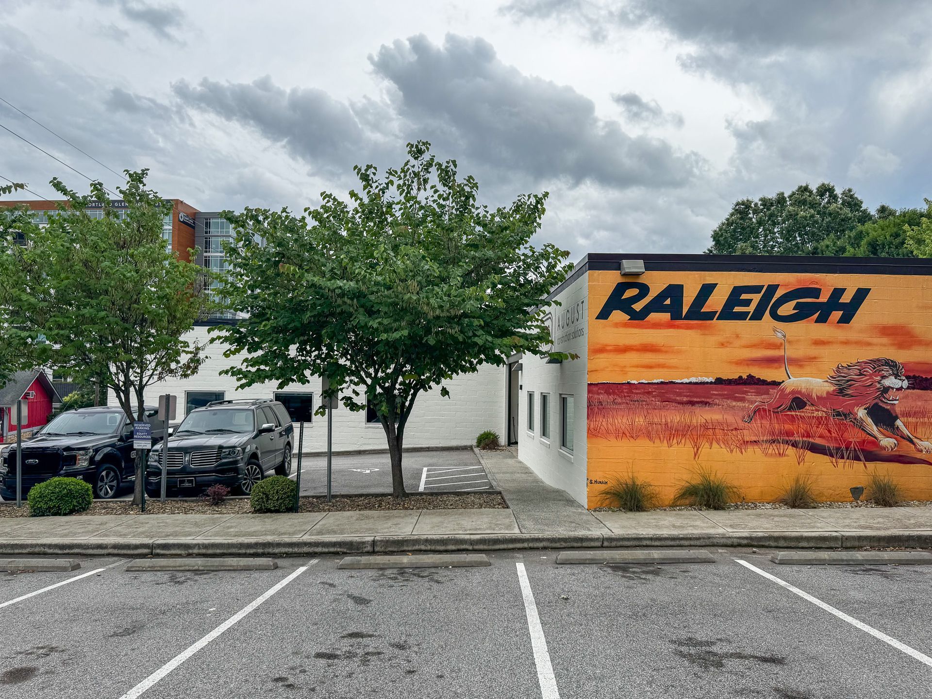 Exterior of Raleigh building with a colorful mural and parked cars. Trees and cloudy sky.