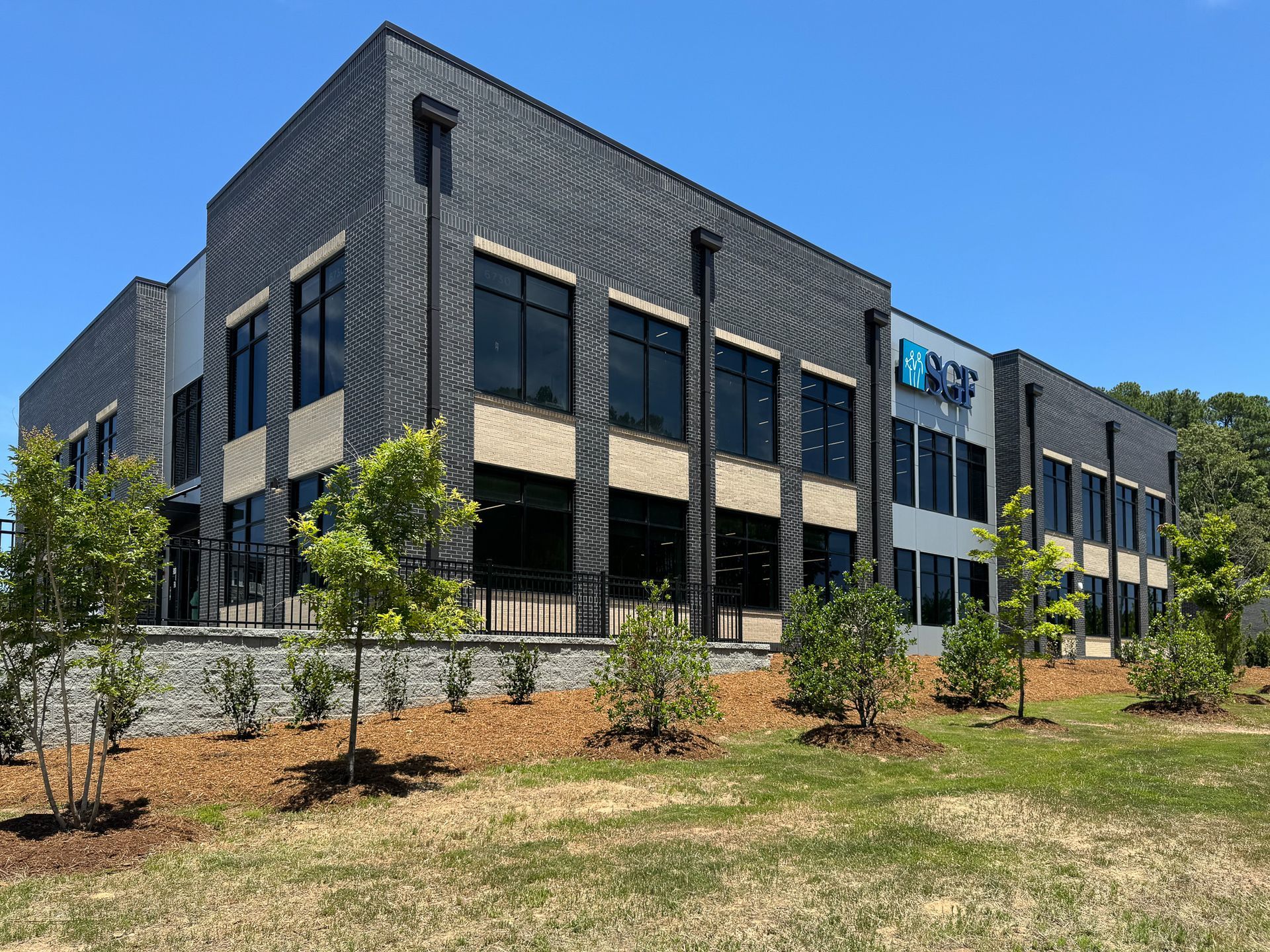 Modern two-story office building with dark brick facade, large windows, and manicured landscaping under a blue sky.
