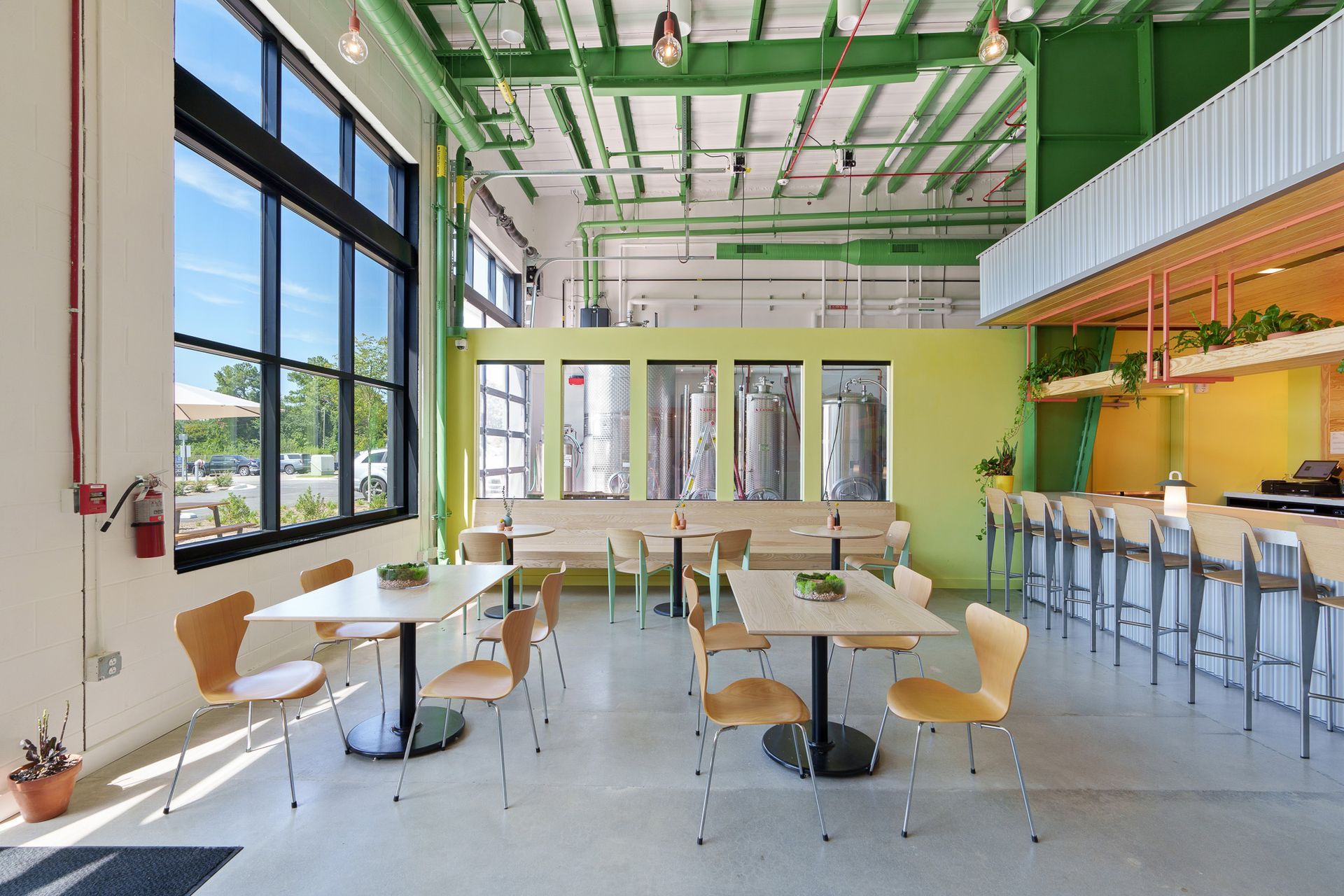 Bright, modern restaurant interior. Tables and chairs in front of large windows, bar in the background. Green and yellow accents.
