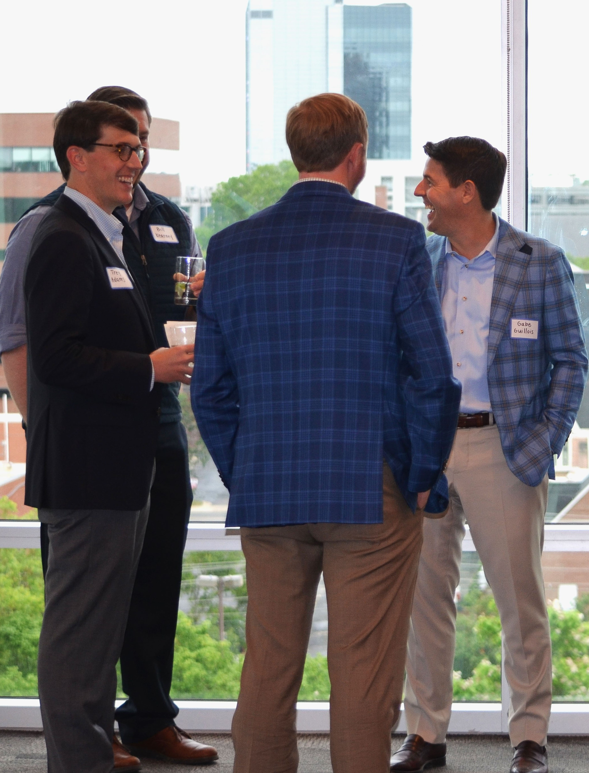 Four men in business attire socializing near a window with city views. Two are laughing.