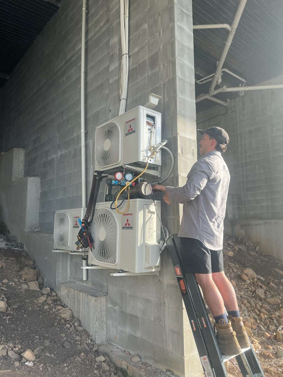 A Man is Standing on a Ladder Working on an Air Conditioner — Kilowatts & Cool Whitsundays In Strathdickie, QLD