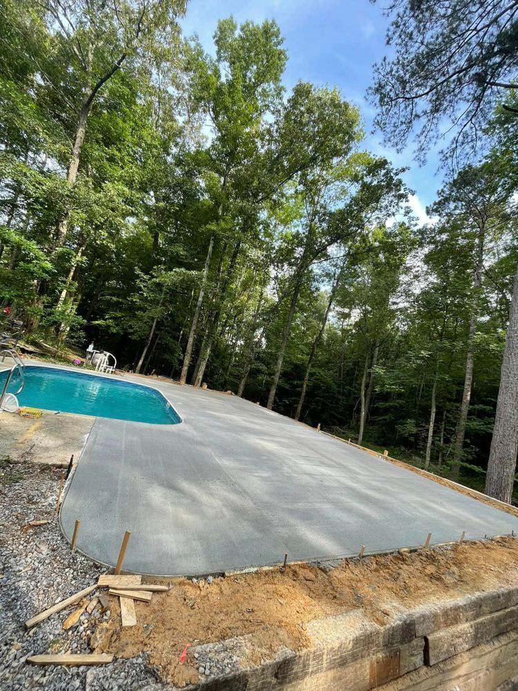 A concrete patio is being built next to a swimming pool surrounded by trees.