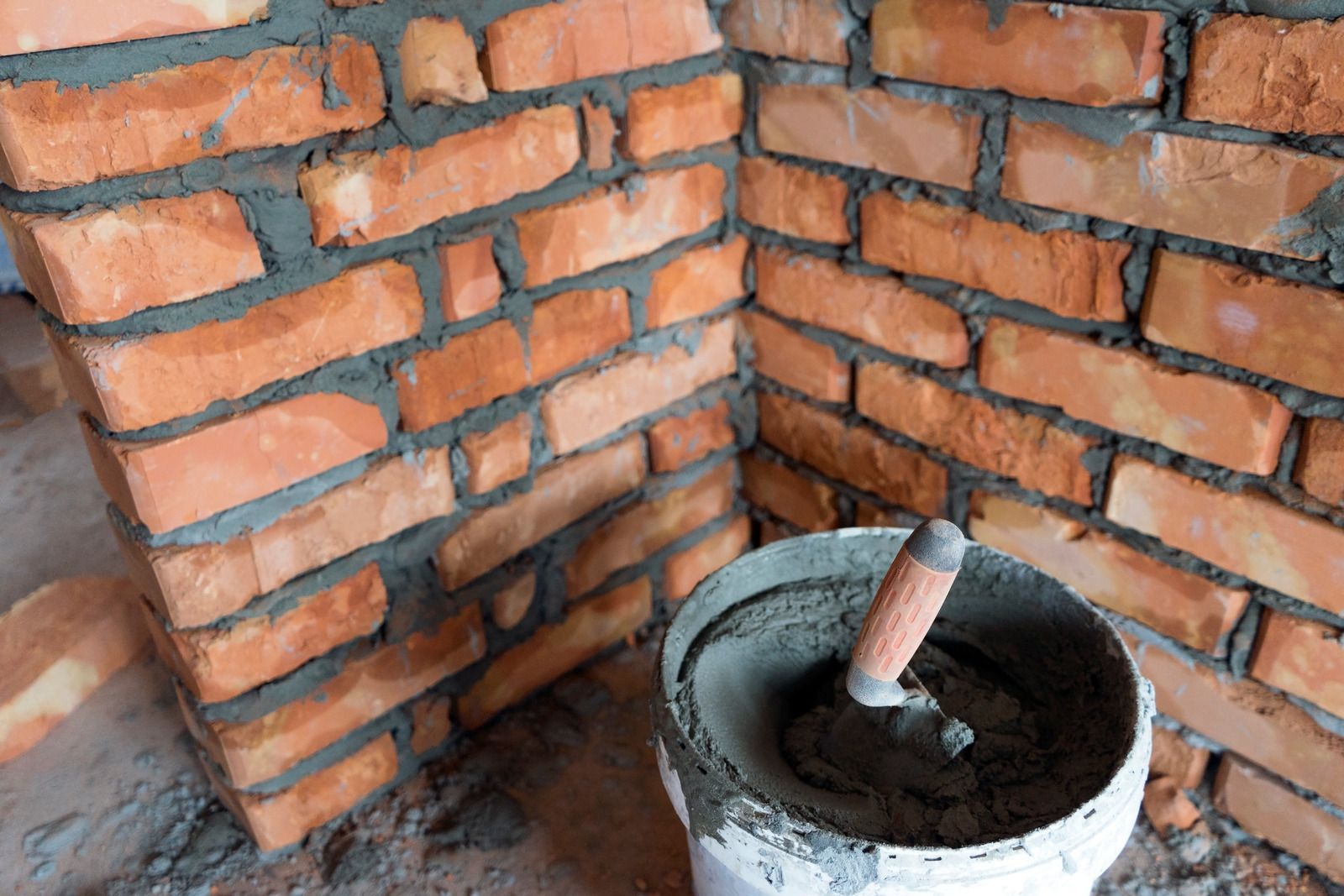 A bucket of cement and a trowel are sitting in front of a brick wall.