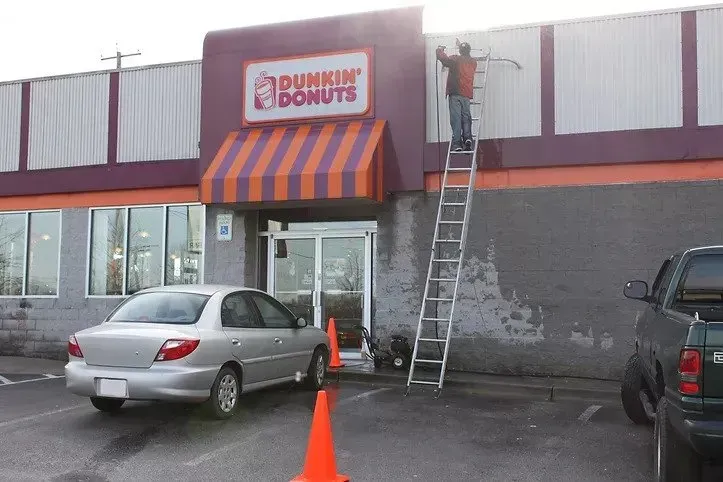 A dunkin donuts store is being painted by a man on a ladder