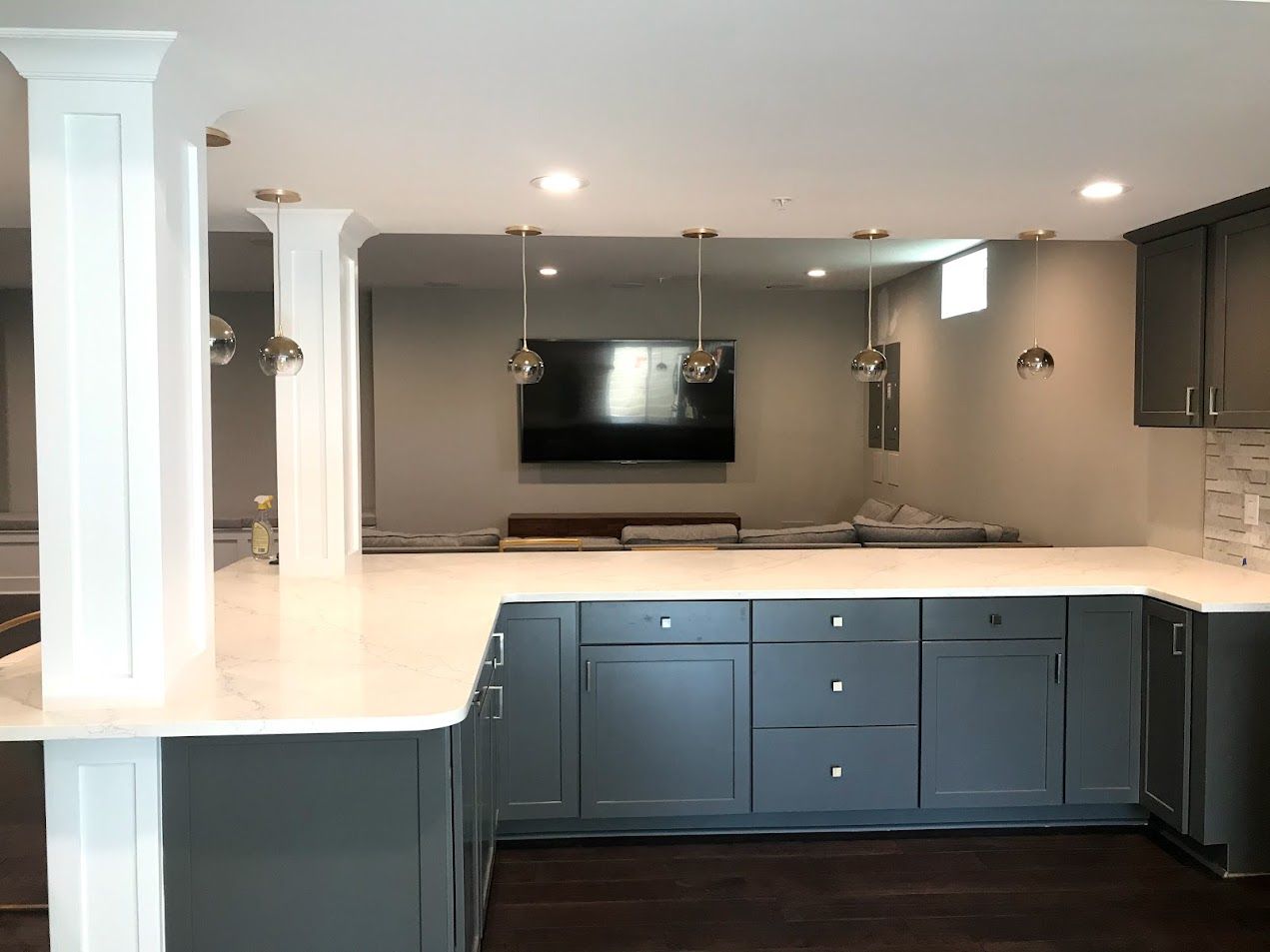 A kitchen with gray cabinets and white counter tops and a flat screen tv on the wall.