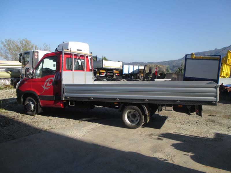 Un camioncino rosso con pianale ribassato parcheggiato all'aperto su un piazzale ghiaioso sotto un cielo azzurro e limpido.