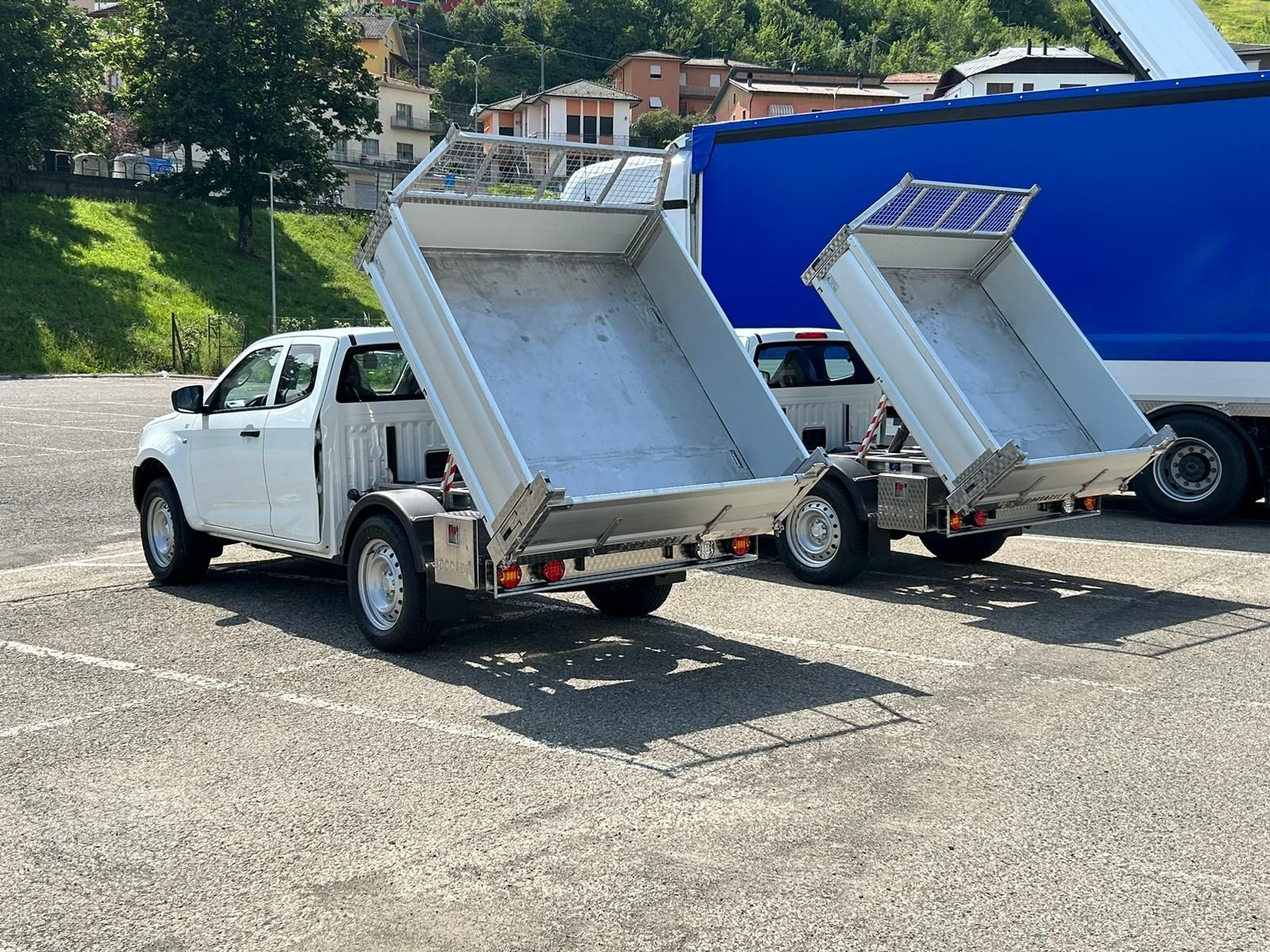 Due pick-up bianchi con il cassone ribaltabile in metallo sollevato, parcheggiati su un piazzale ghiaioso di fronte a un camion blu.