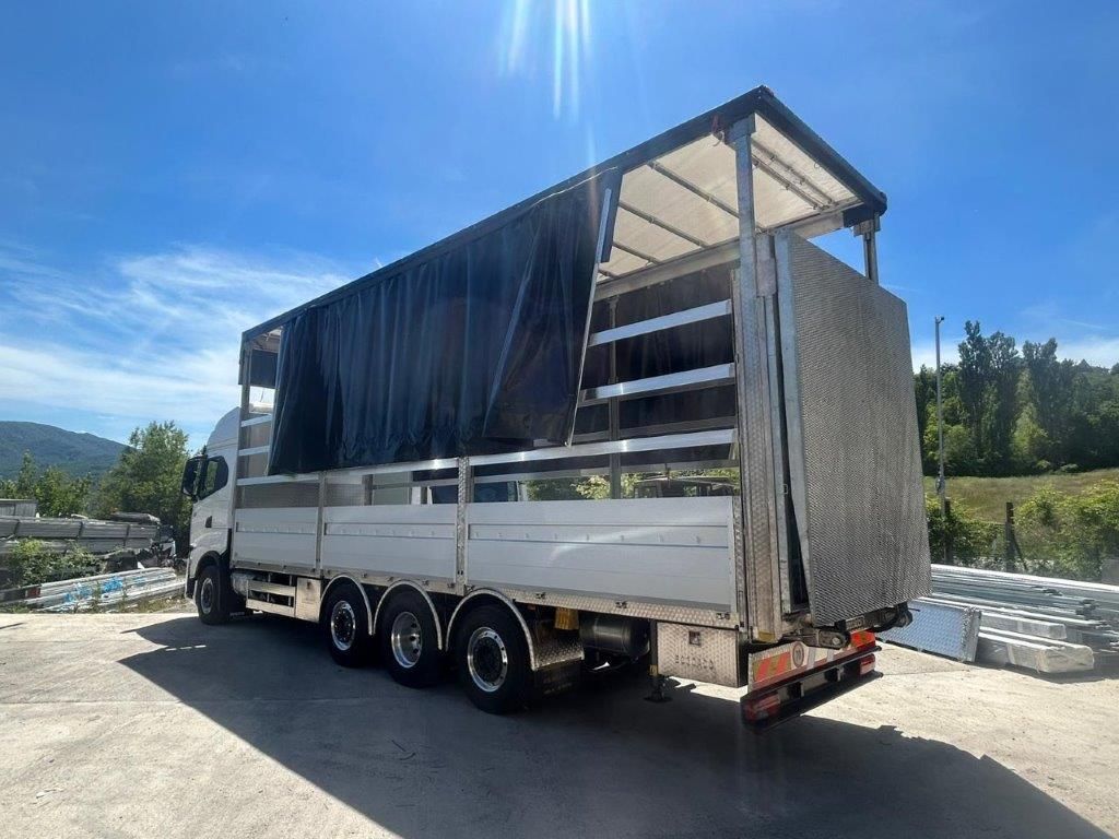 Un camioncino bianco con pianale e telone laterale nero parzialmente aperto, parcheggiato in un parcheggio all'aperto soleggiato in campagna.