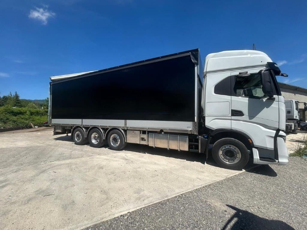 Un camioncino bianco con un lungo rimorchio coperto di nero parcheggiato su un piazzale ghiaioso sotto un cielo azzurro brillante.