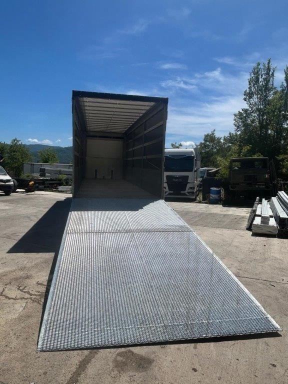 La parte posteriore aperta di un grande autoarticolato parcheggiato all'aperto, con un'ampia rampa metallica che conduce al vano di carico.