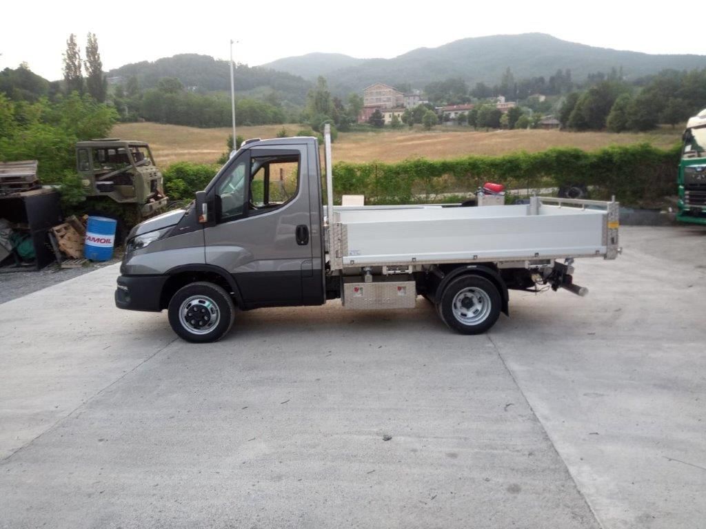 Un camioncino grigio a cabina singola con pianale ribassato parcheggiato su un piazzale di cemento, con un paesaggio collinare ed erboso sullo sfondo.
