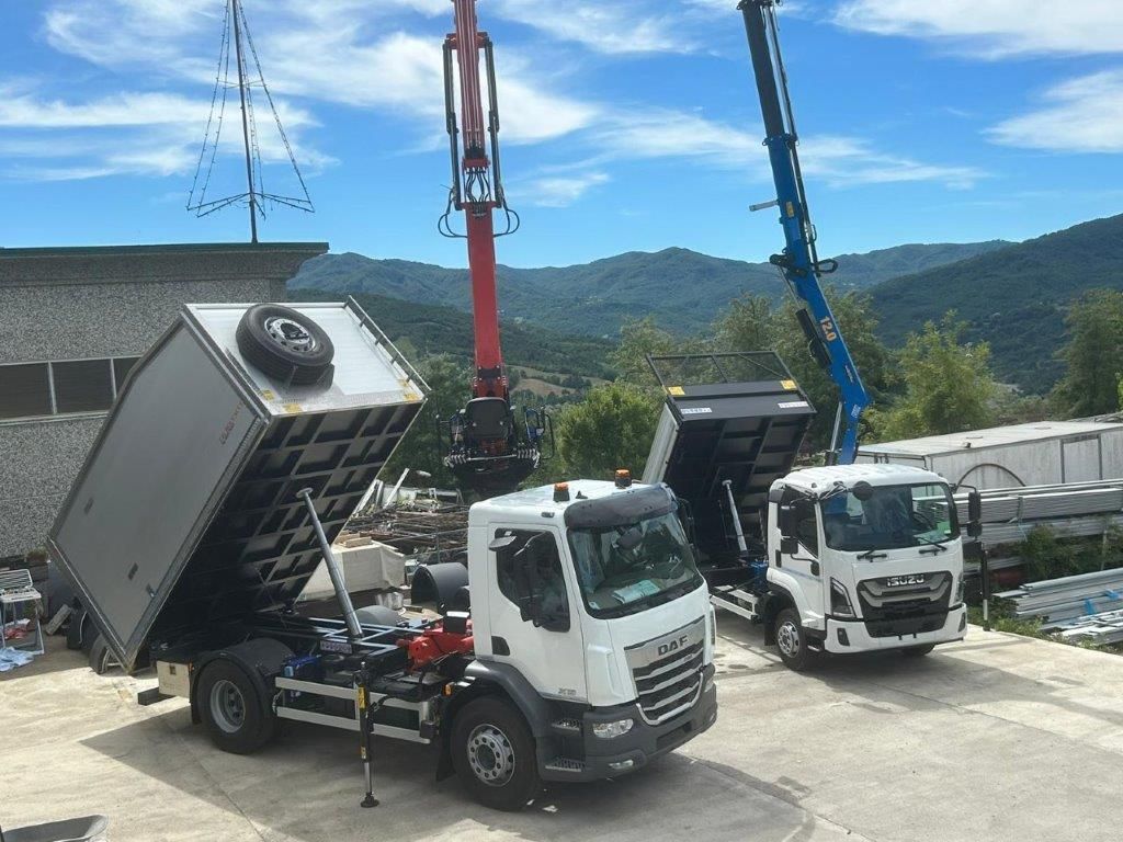 Due autocarri bianchi per carichi pesanti con cassone ribaltabile rialzato, parcheggiati fianco a fianco con delle gru in un soleggiato piazzale industriale.