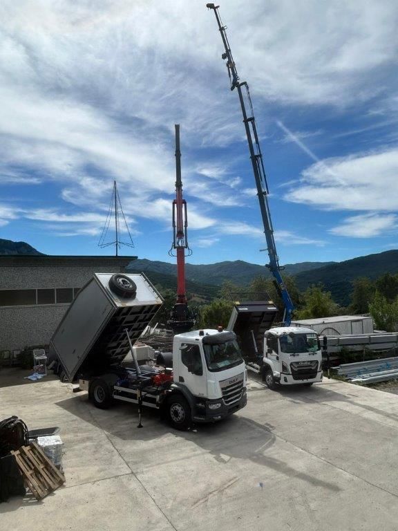 Due autocarri ribaltabili bianchi parcheggiati su un piazzale di cemento, uno con il cassone sollevato, accanto a un'alta gru sullo sfondo di un panorama montano.