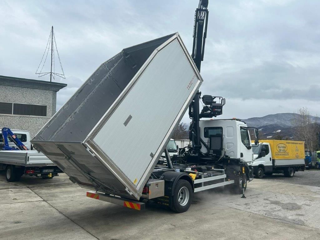 Un camion bianco con una gru montata a bordo inclina un grande container metallico vuoto su un piazzale asfaltato.
