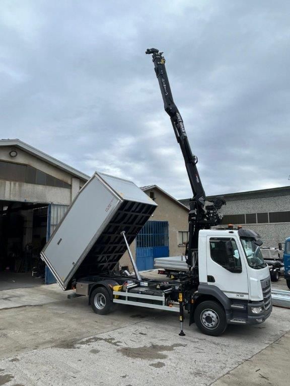 Un camion bianco con un braccio gru estensibile e un pianale di carico rialzato è parcheggiato su un piazzale asfaltato di fronte a un edificio.