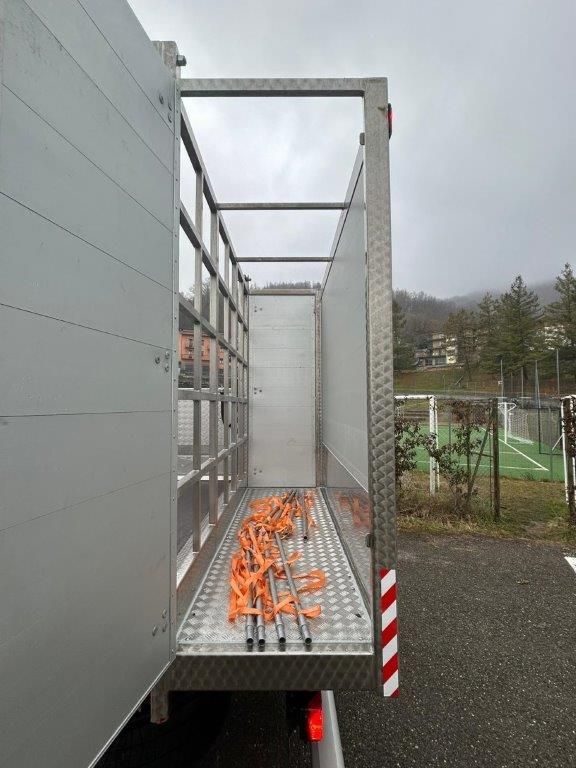Un rimorchio multiuso aperto sui lati, con telaio e pianale in metallo, contenente cinghie di fissaggio arancioni, su un piazzale asfaltato.