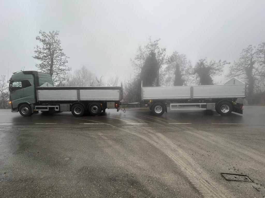 Un autocarro ribaltabile argentato con rimorchio, parcheggiato su una superficie asfaltata, sullo sfondo di alberi avvolti da una fitta nebbia grigia.