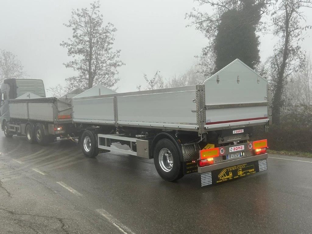 Vista posteriore di un rimorchio a pianale nero con rampe di carico metalliche verticali, parcheggiato in un'area industriale all'aperto.