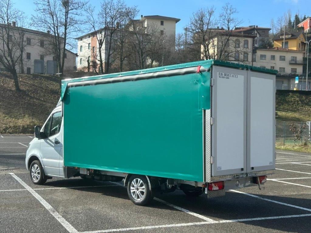 Un camion bianco con un grande container verde per il carico parcheggiato in un piazzale asfaltato vuoto vicino ad alcuni edifici in una giornata di sole.