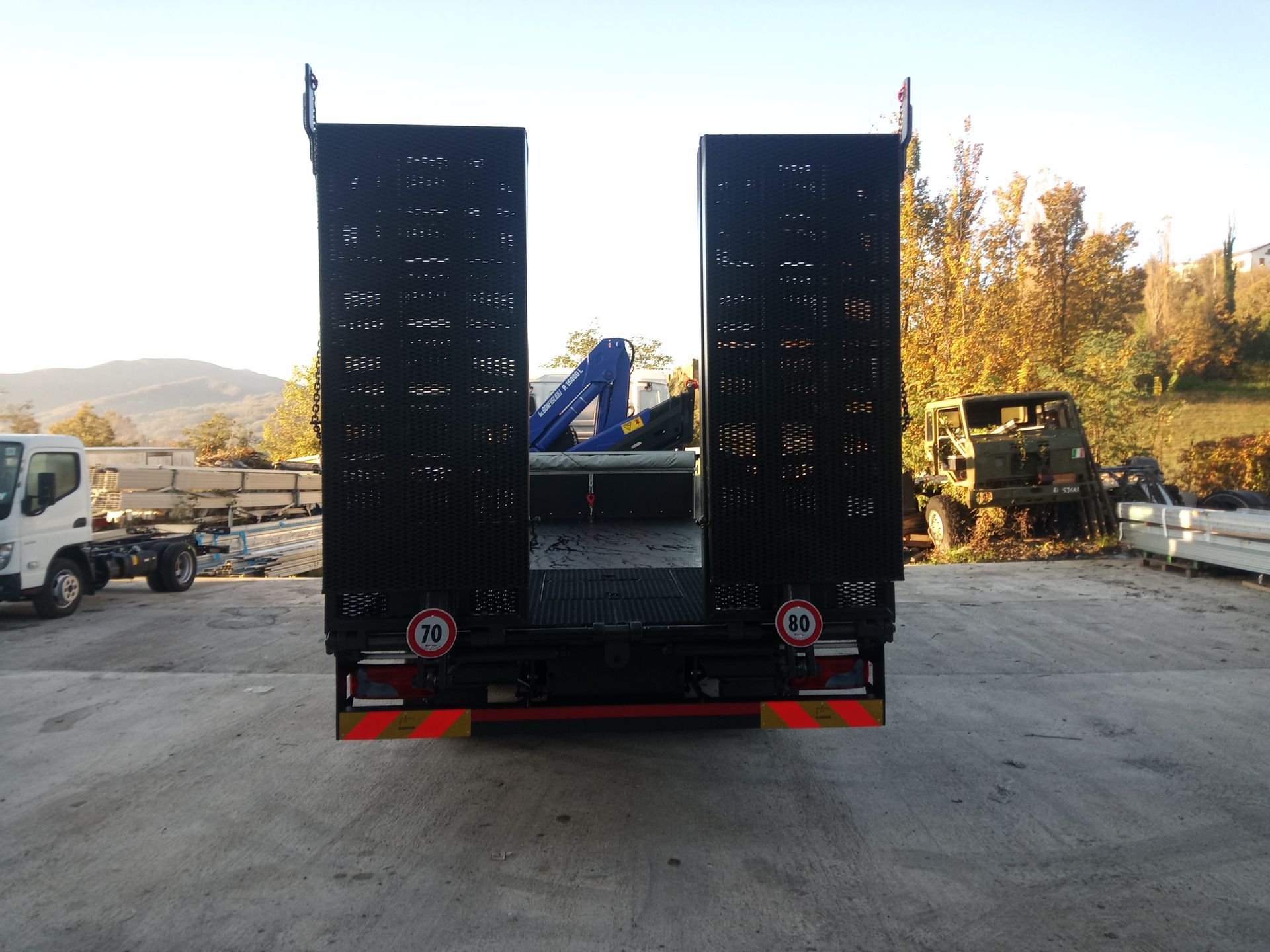 Vista posteriore di un rimorchio a pianale nero con rampe di carico metalliche verticali, parcheggiato in un'area industriale all'aperto.