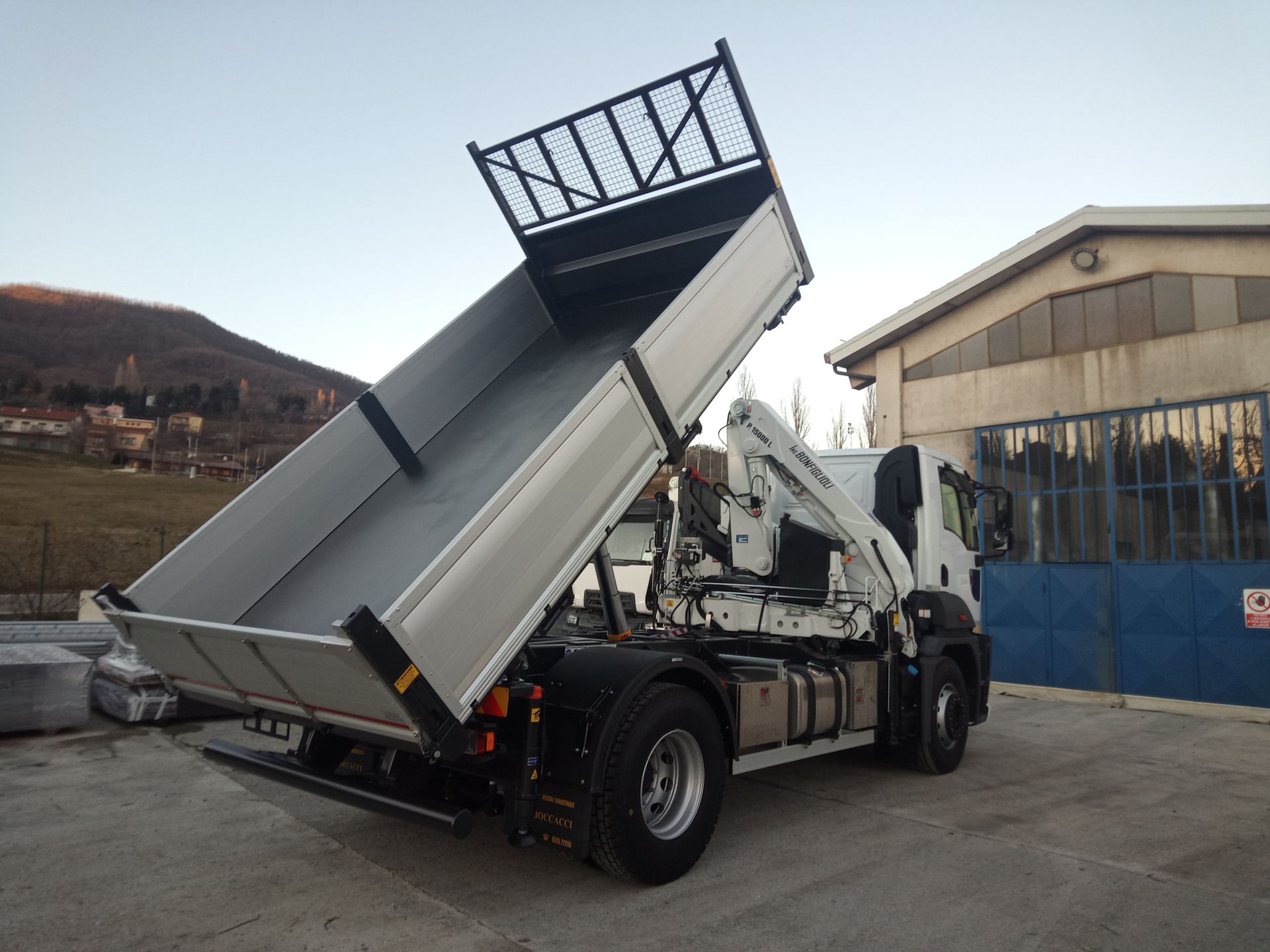 Un autocarro ribaltabile bianco con il cassone idraulico sollevato, parcheggiato in un'area industriale con un edificio sullo sfondo.