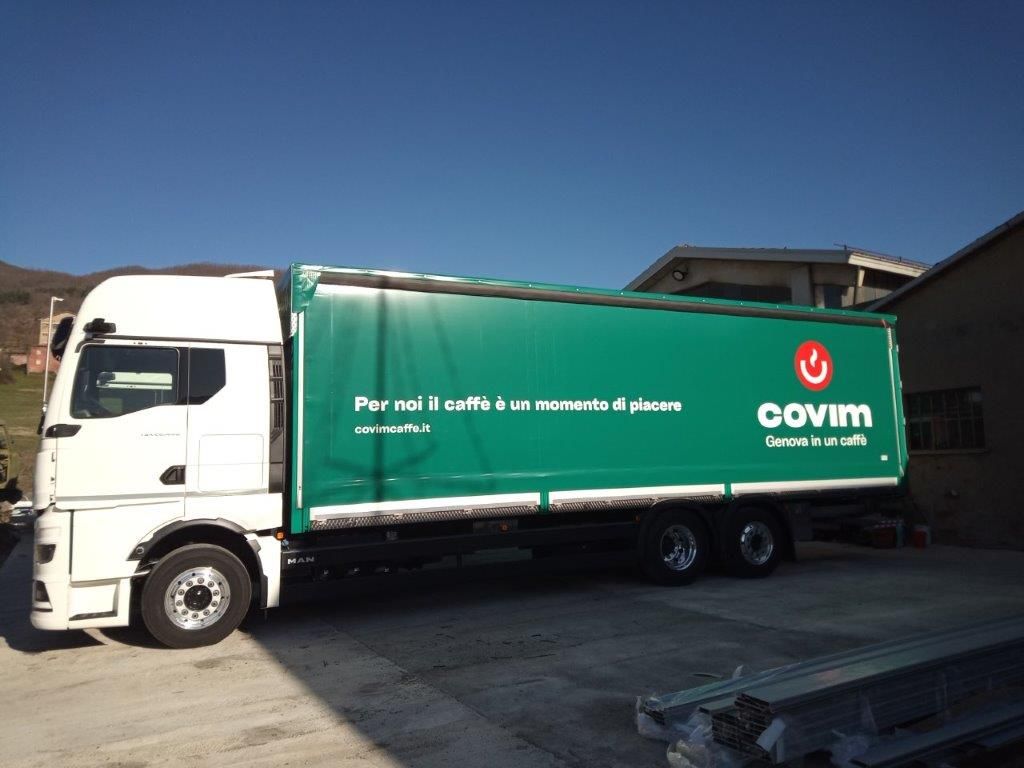 Un semirimorchio bianco con un grande rimorchio verde su cui è stampato il logo del caffè Govim, parcheggiato su un piazzale di cemento all'aperto.