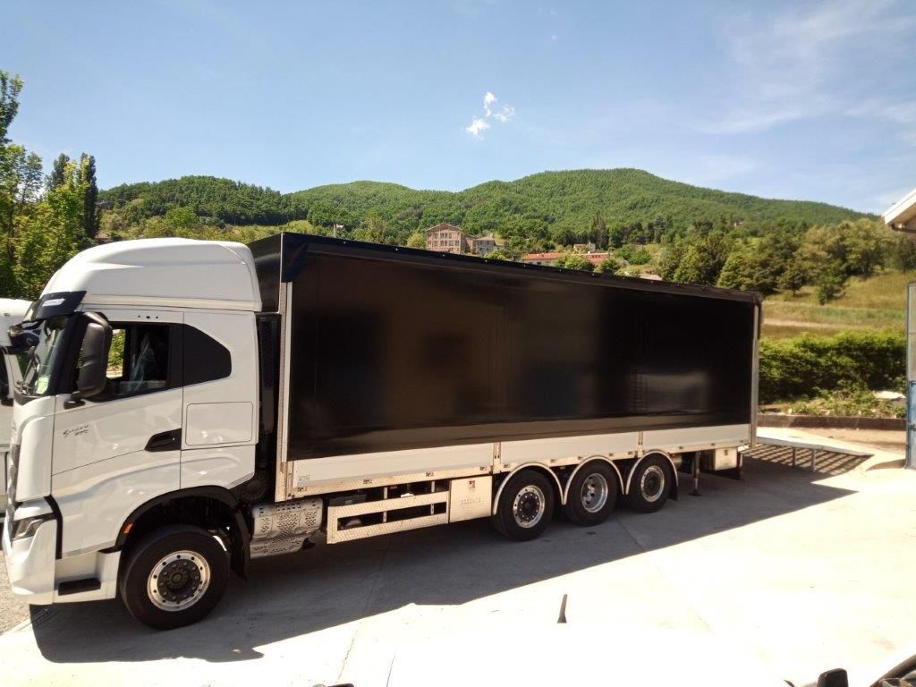 Un camion bianco con un grande rimorchio nero per il carico, parcheggiato su una superficie di cemento, sullo sfondo di colline e cielo azzurro.