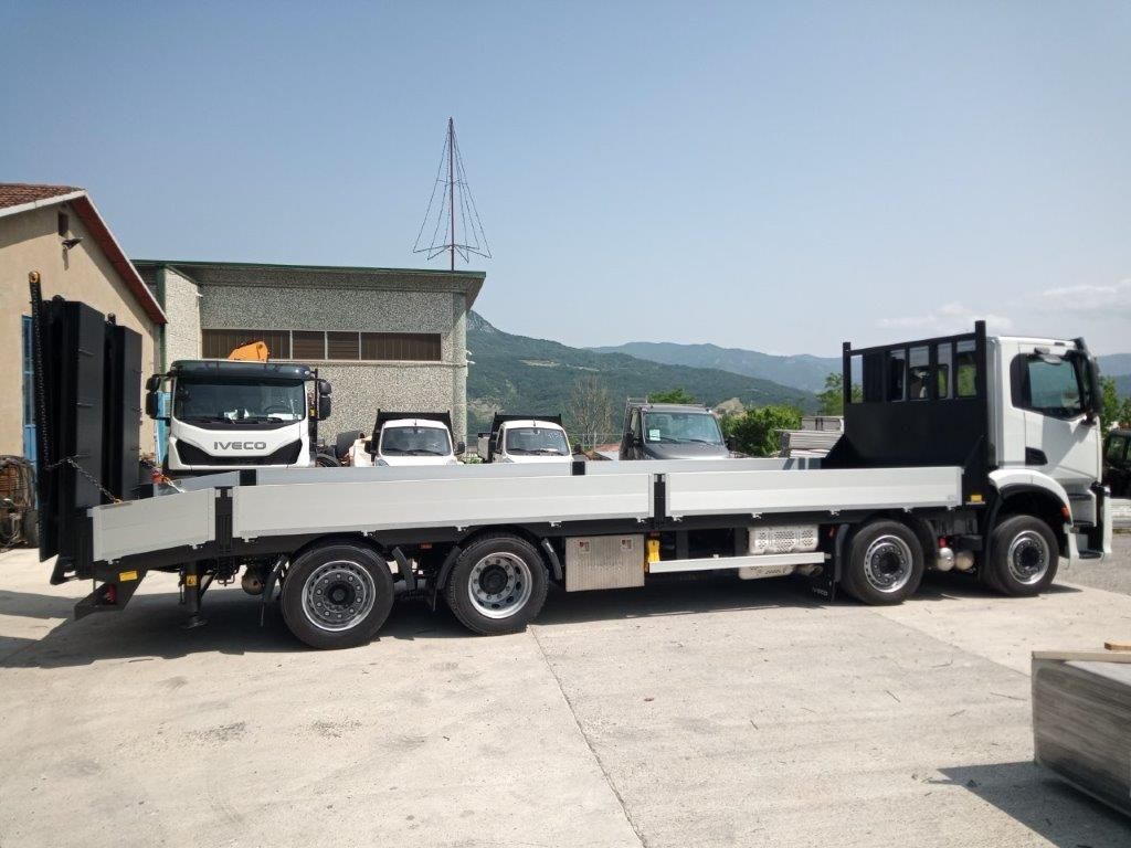 Un camioncino bianco con pianale e rampa di carico posteriore parcheggiato in un piazzale esterno asfaltato sotto un cielo azzurro e limpido.