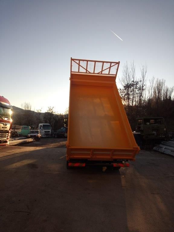 Il cassone di un camion ribaltabile giallo, sollevato e inclinato, è parcheggiato in un piazzale al tramonto.