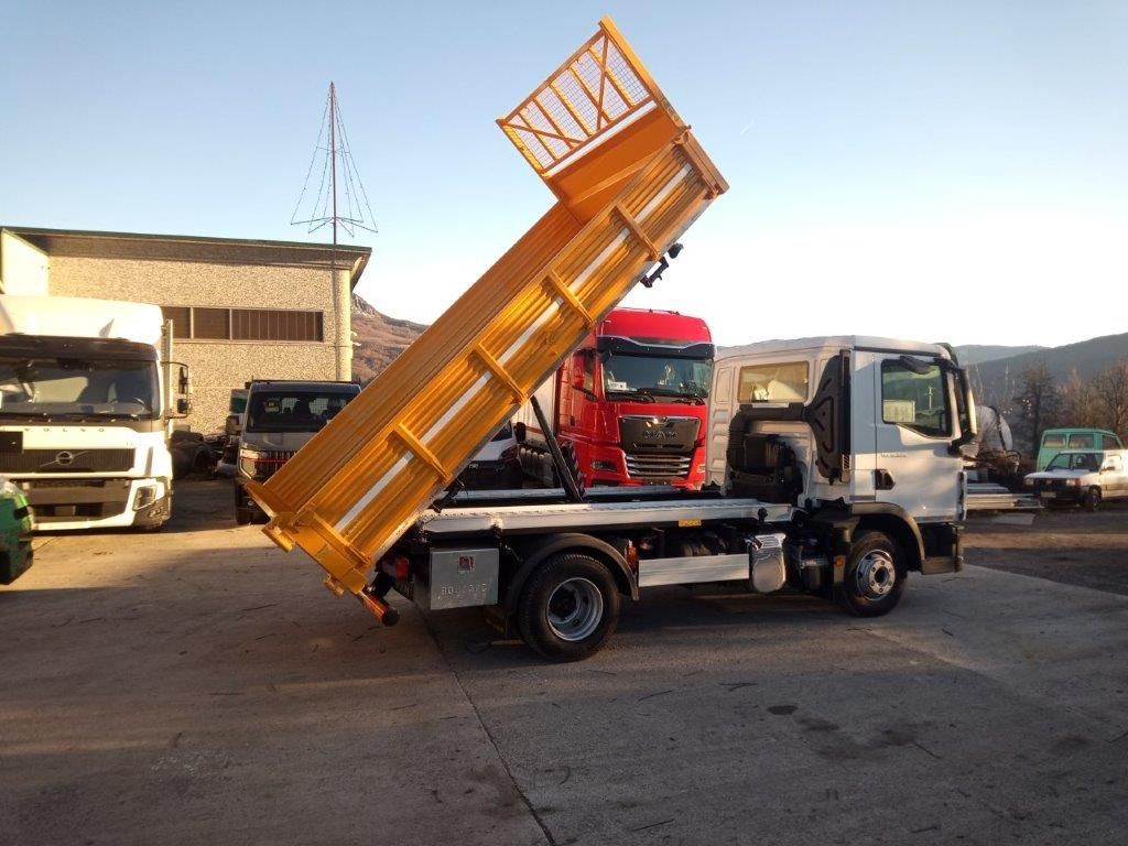 Un autocarro ribaltabile bianco con il cassone giallo sollevato, parcheggiato in un piazzale con altri camion al tramonto.