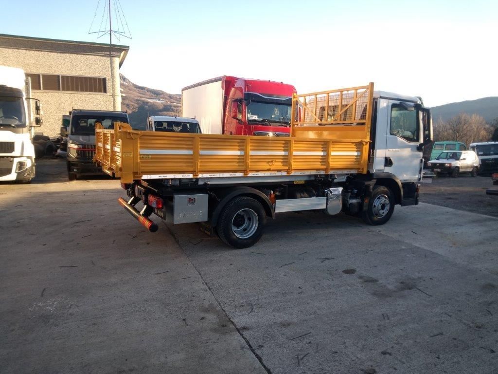 Un camion bianco con un pianale di carico giallo è parcheggiato in un piazzale ghiaioso, con altri veicoli visibili sullo sfondo.
