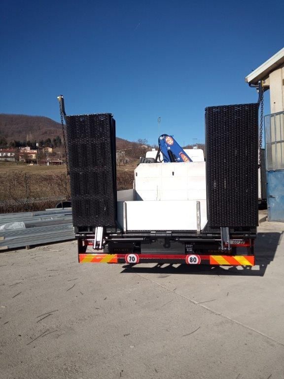 Vista posteriore di un rimorchio a pianale con rampe di carico verticali in metallo nero su un piazzale ghiaioso sotto un cielo azzurro e limpido.