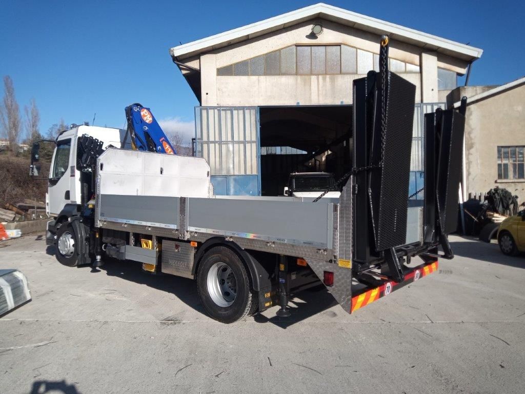Un camioncino bianco con pianale, dotato di una gru blu montata dietro la cabina e rampe di carico verticali, parcheggiato all'esterno di un magazzino.