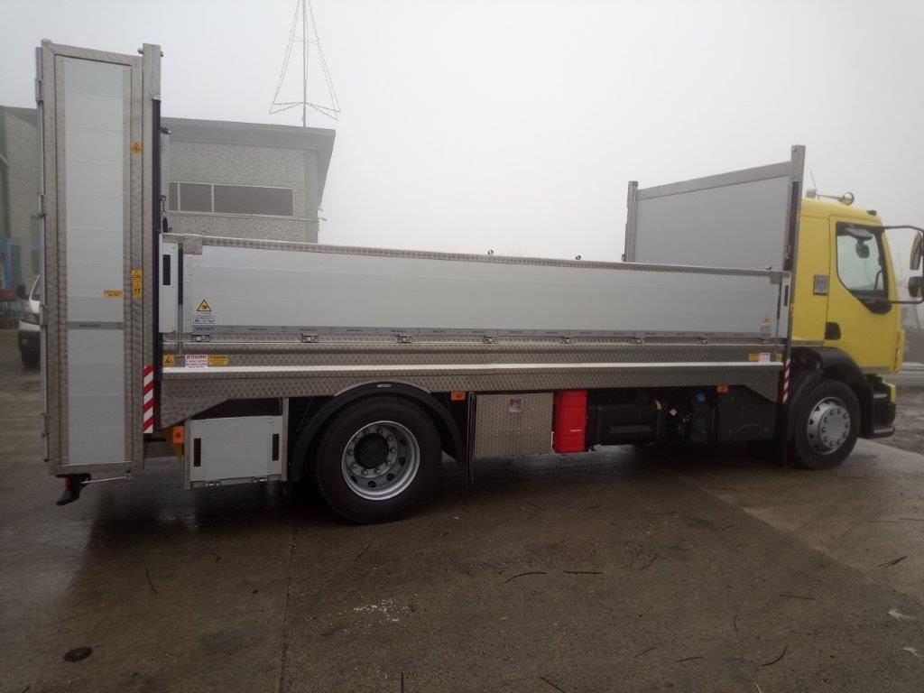 Un camioncino giallo con pianale, pannelli laterali argentati e sponda posteriore verticale, parcheggiato in una giornata grigia e nuvolosa.