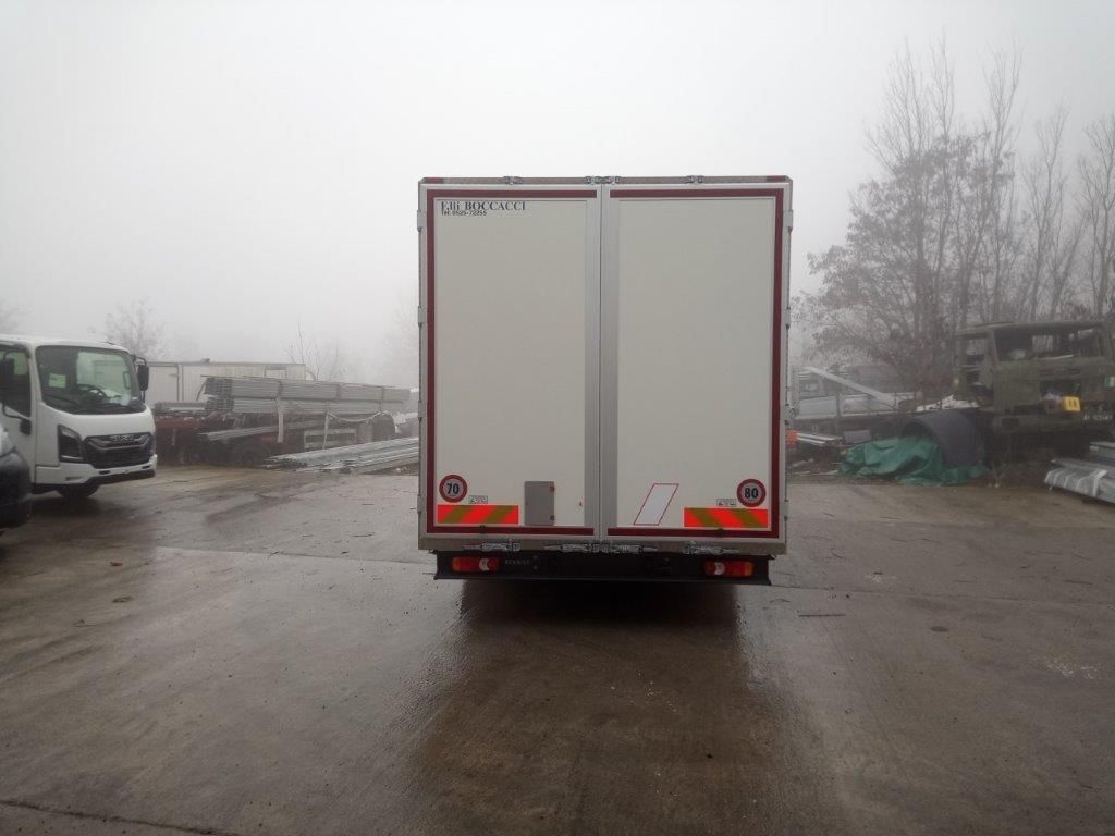 Una gru rossa montata su un camion, ripiegata per il trasporto, è parcheggiata in un piazzale industriale innevato vicino ad alcuni edifici.