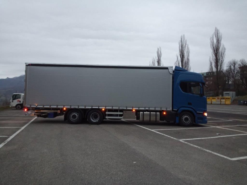 Un camion blu con un lungo rimorchio grigio parcheggiato in un piazzale asfaltato sotto un cielo nuvoloso.