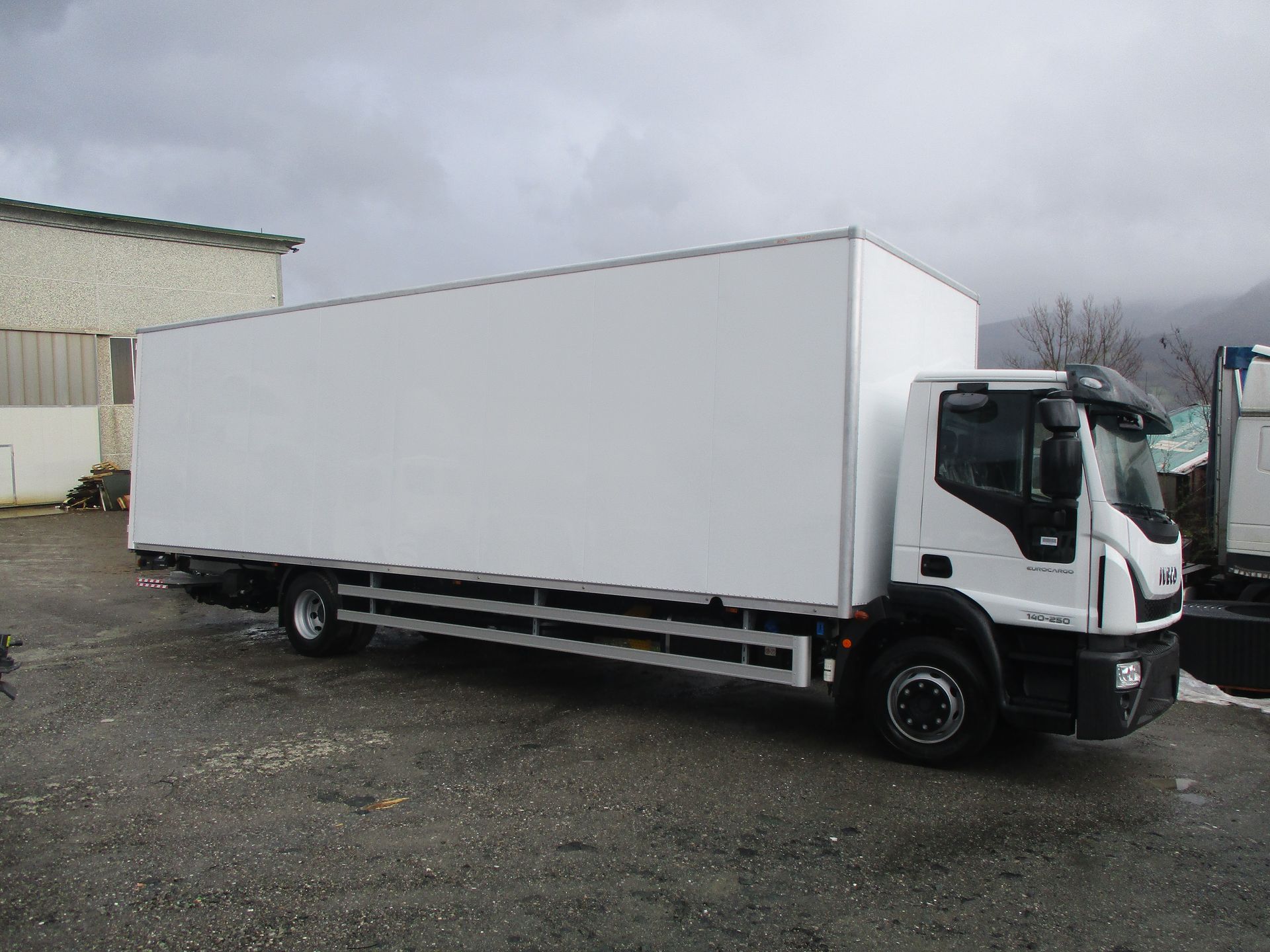 Un furgone bianco parcheggiato su un piazzale ghiaioso di fronte a un edificio, sotto un cielo nuvoloso.
