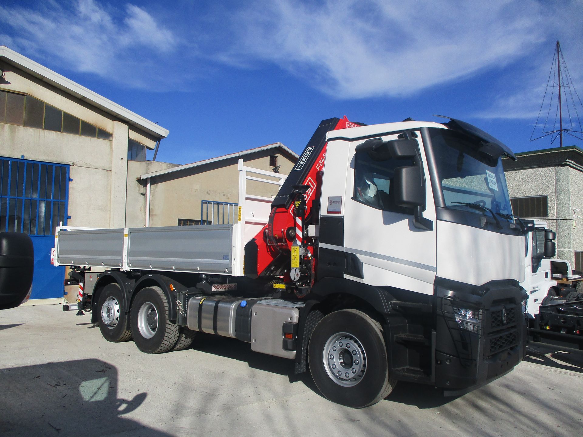 Un autocarro ribaltabile Renault bianco con una gru rossa montata sul telaio, parcheggiato su un piazzale asfaltato di fronte a degli edifici industriali.