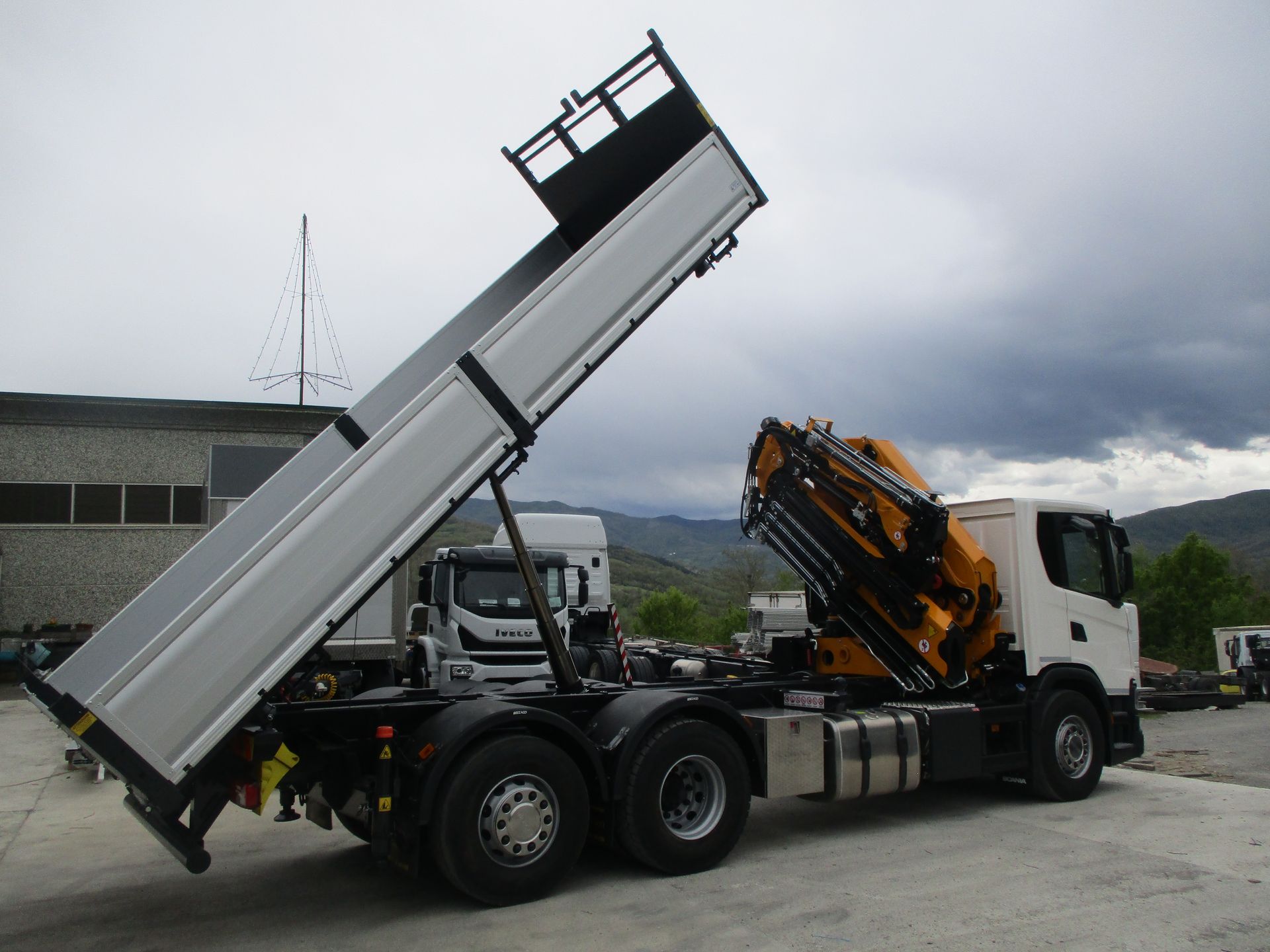 Un autocarro ribaltabile bianco con un braccio della gru giallo, parcheggiato in un piazzale con il cassone ribaltabile sollevato in aria.