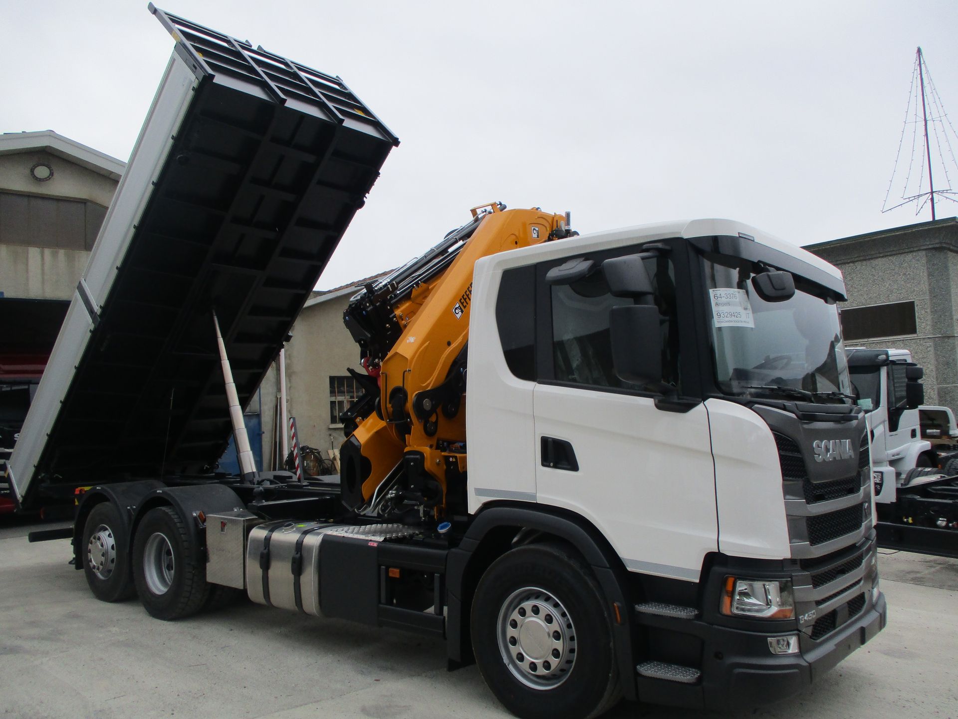 Un autocarro ribaltabile Scania bianco con gru idraulica gialla e cassone di carico aperto e rialzato, parcheggiato in un piazzale.