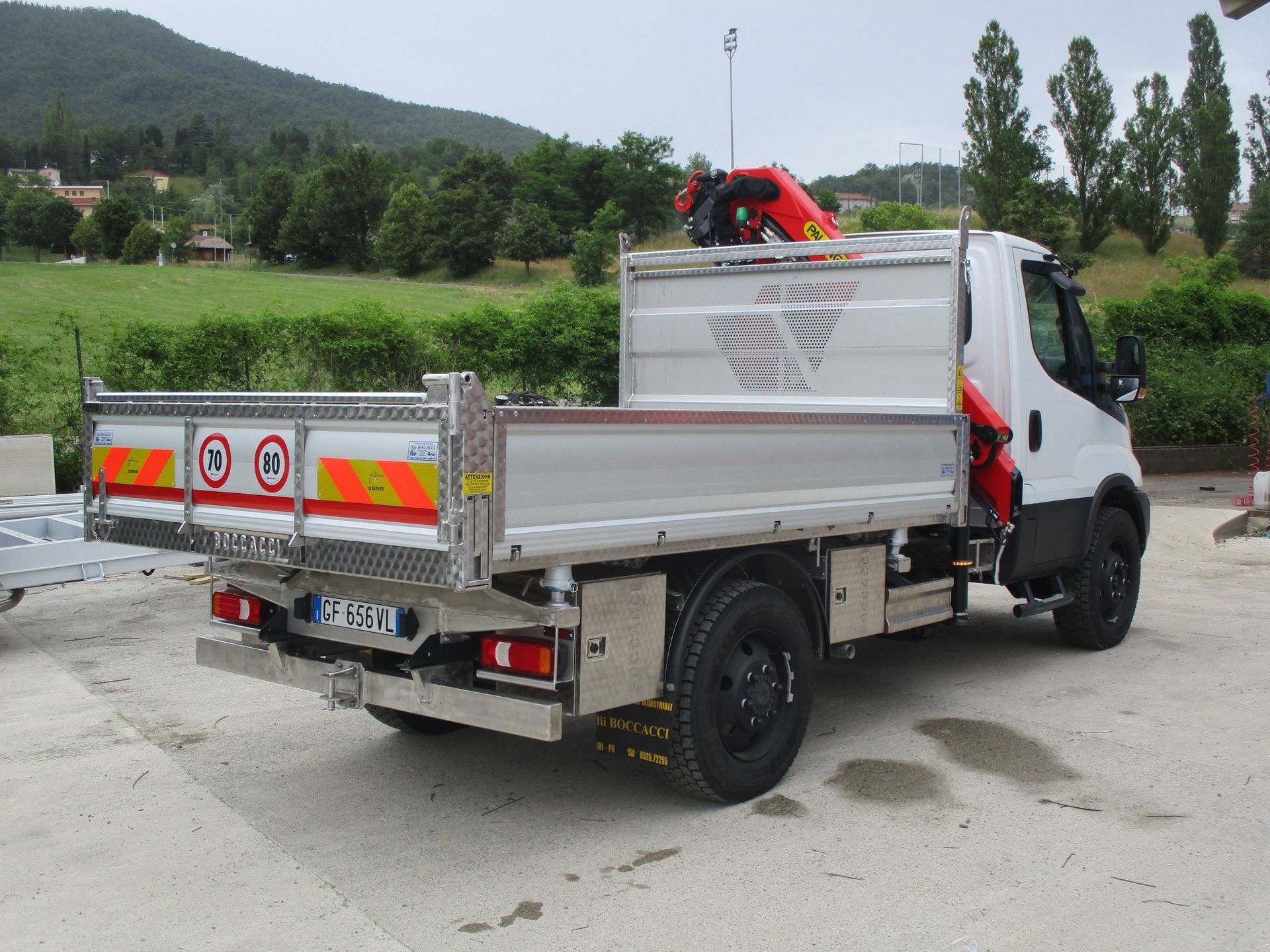 Un camion bianco a pianale ribassato con gru agganciata, parcheggiato su un piazzale asfaltato di fronte a una collina erbosa.