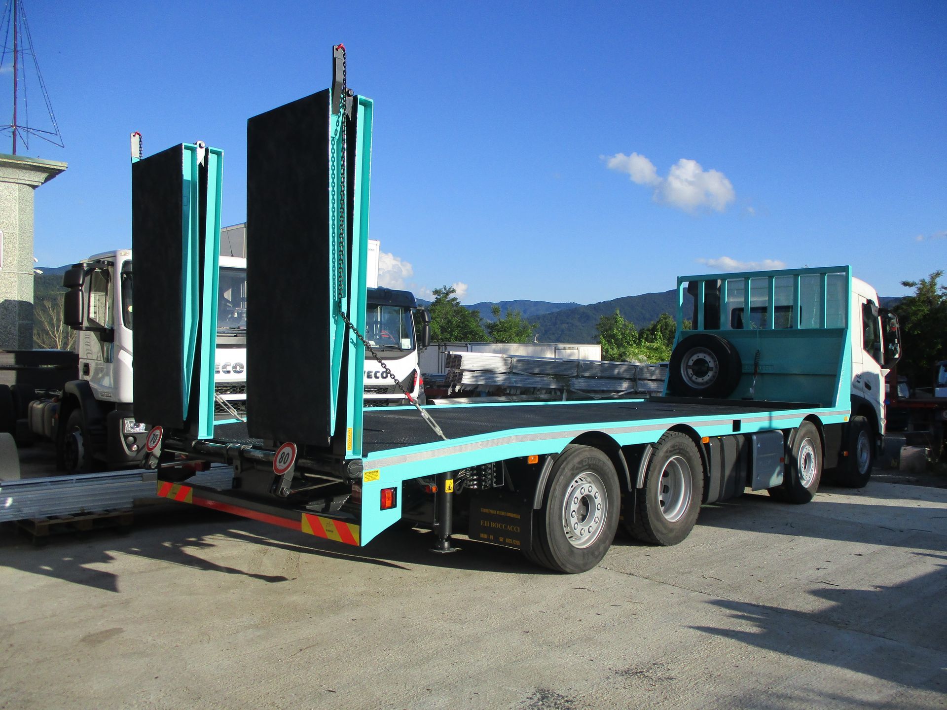 Un camion a pianale di un vivace colore verde acqua, dotato di ampie rampe di carico posteriori, parcheggiato in un piazzale all'aperto in una giornata di sole.
