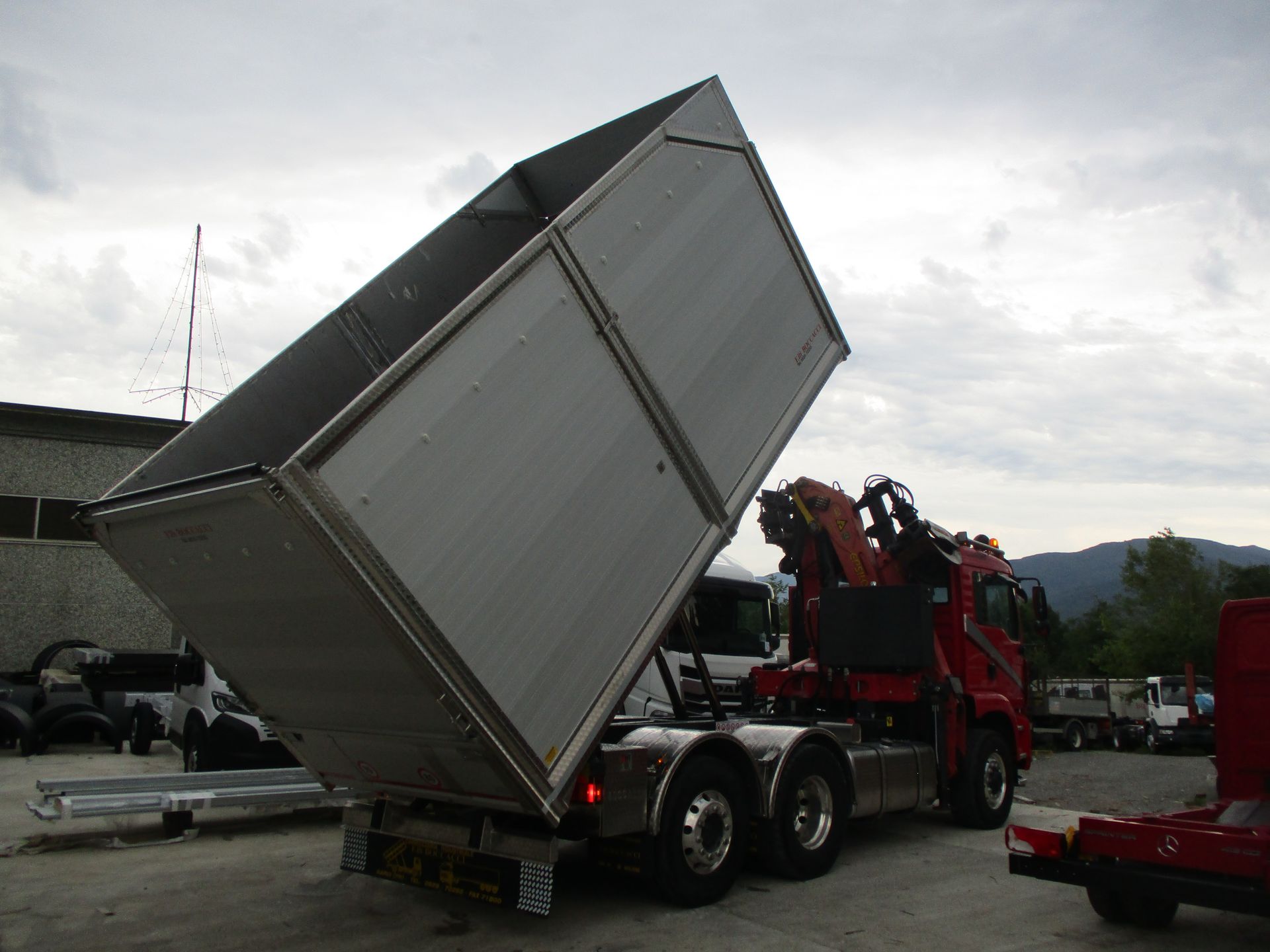 Un camion ribaltabile rosso con un grande cassone metallico vuoto sollevato in un piazzale ghiaioso sotto un cielo nuvoloso.