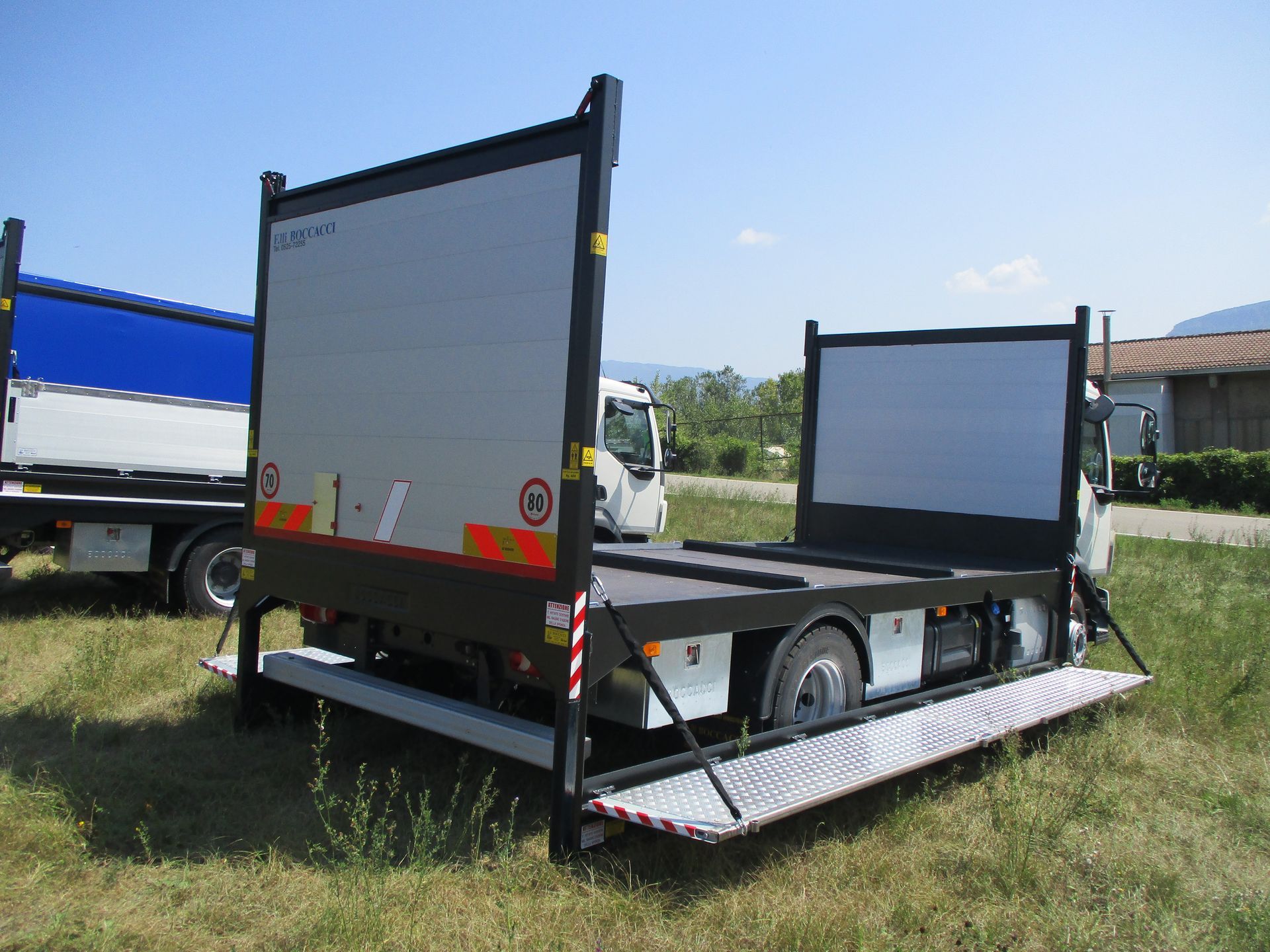 Un camion in un campo con una piattaforma laterale metallica estesa e due pannelli verticali, con un altro camion parcheggiato nelle vicinanze.
