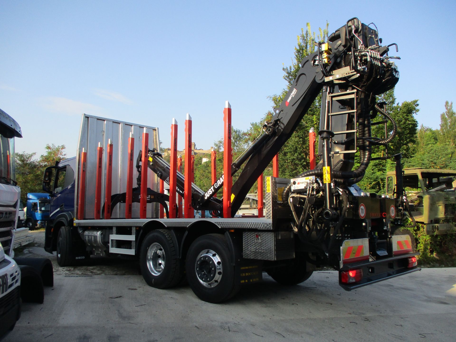 Un camion blu per il trasporto di legname, con una gru idraulica ripiegabile e dei paletti rossi, parcheggiato in un piazzale sotto un cielo sereno.