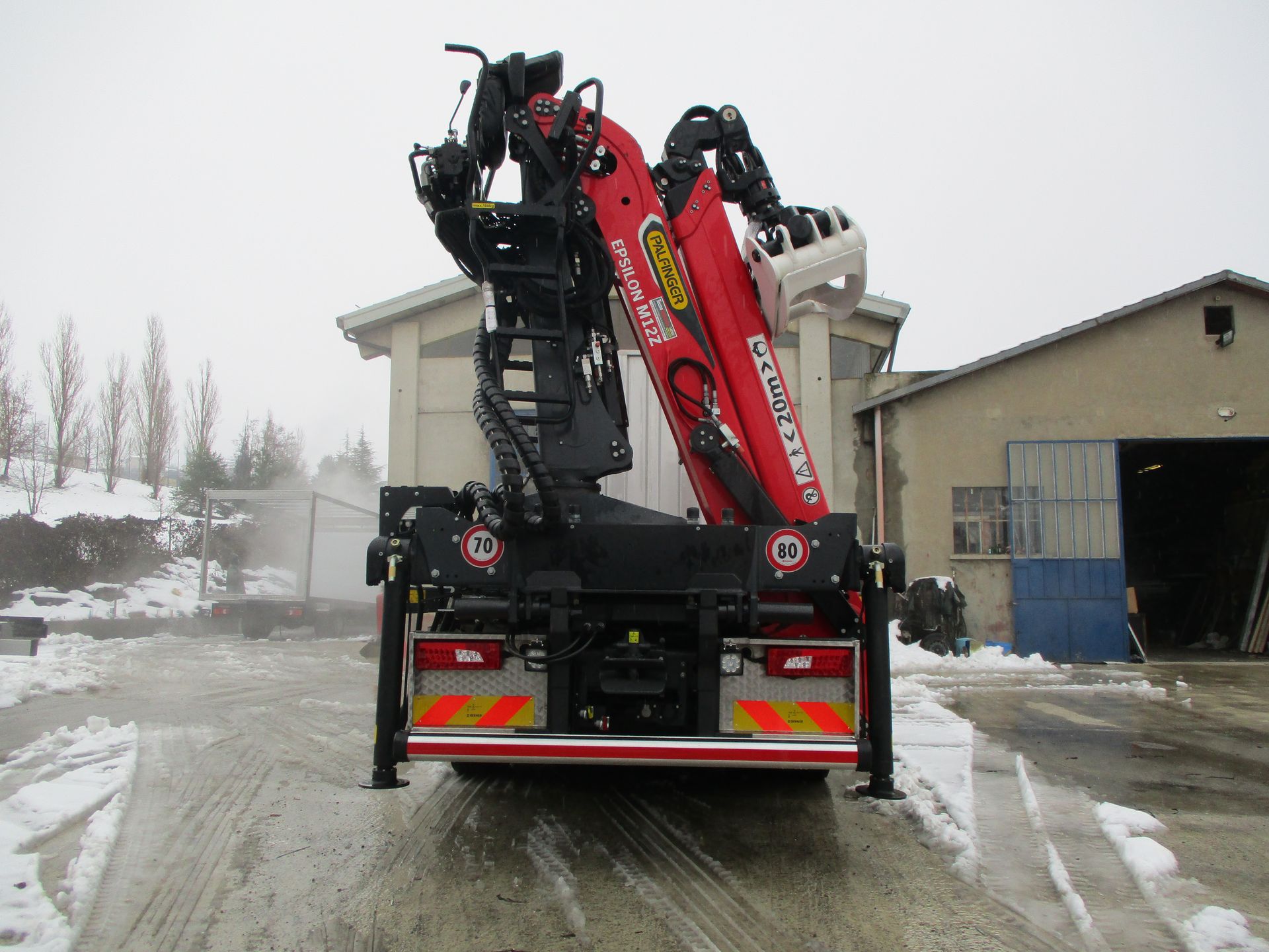 Una gru rossa montata su un camion, ripiegata per il trasporto, è parcheggiata in un piazzale industriale innevato vicino ad alcuni edifici.