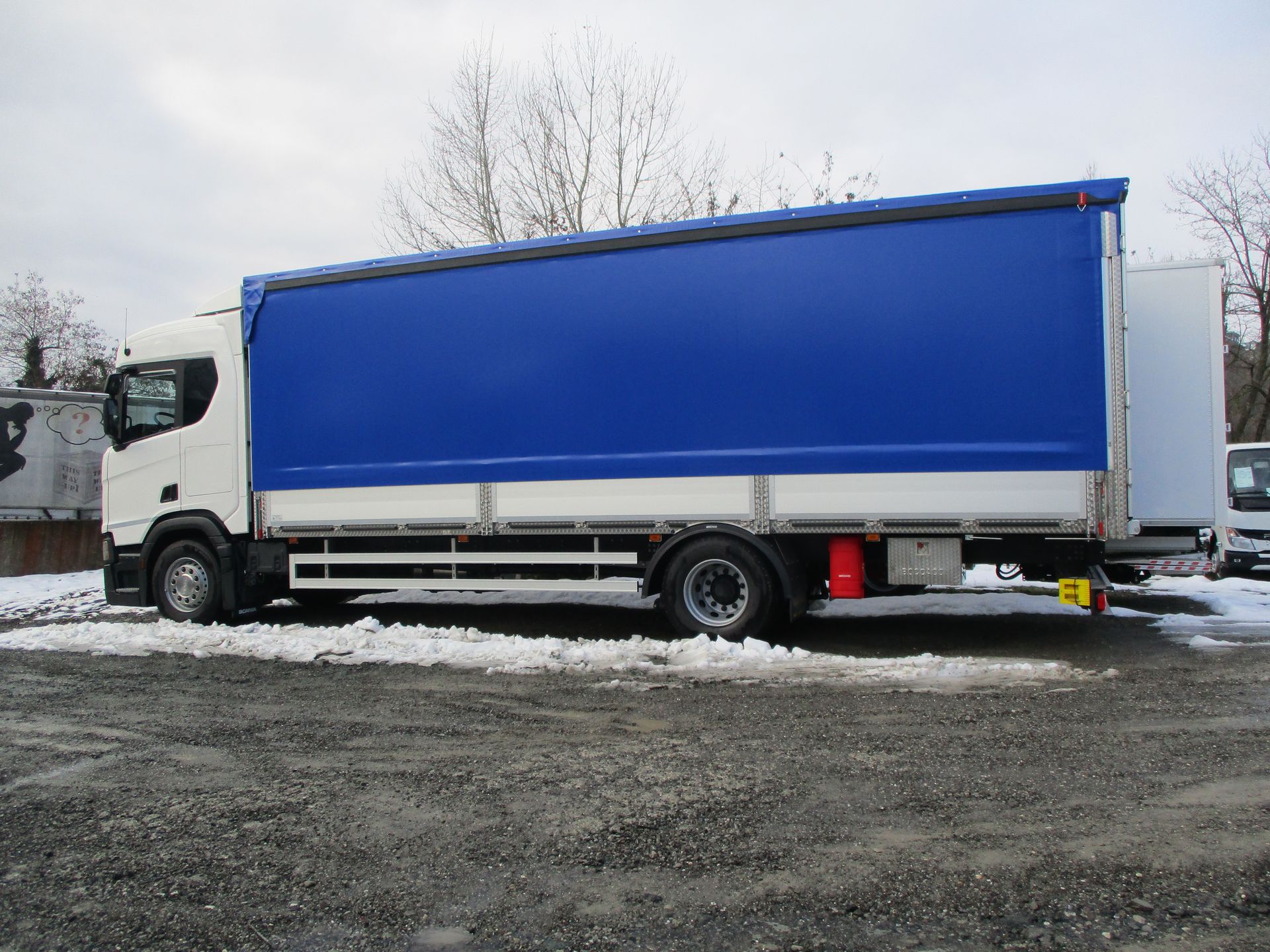 Un camion bianco con un grande rimorchio telonato blu è parcheggiato su un piazzale ghiaioso e innevato.