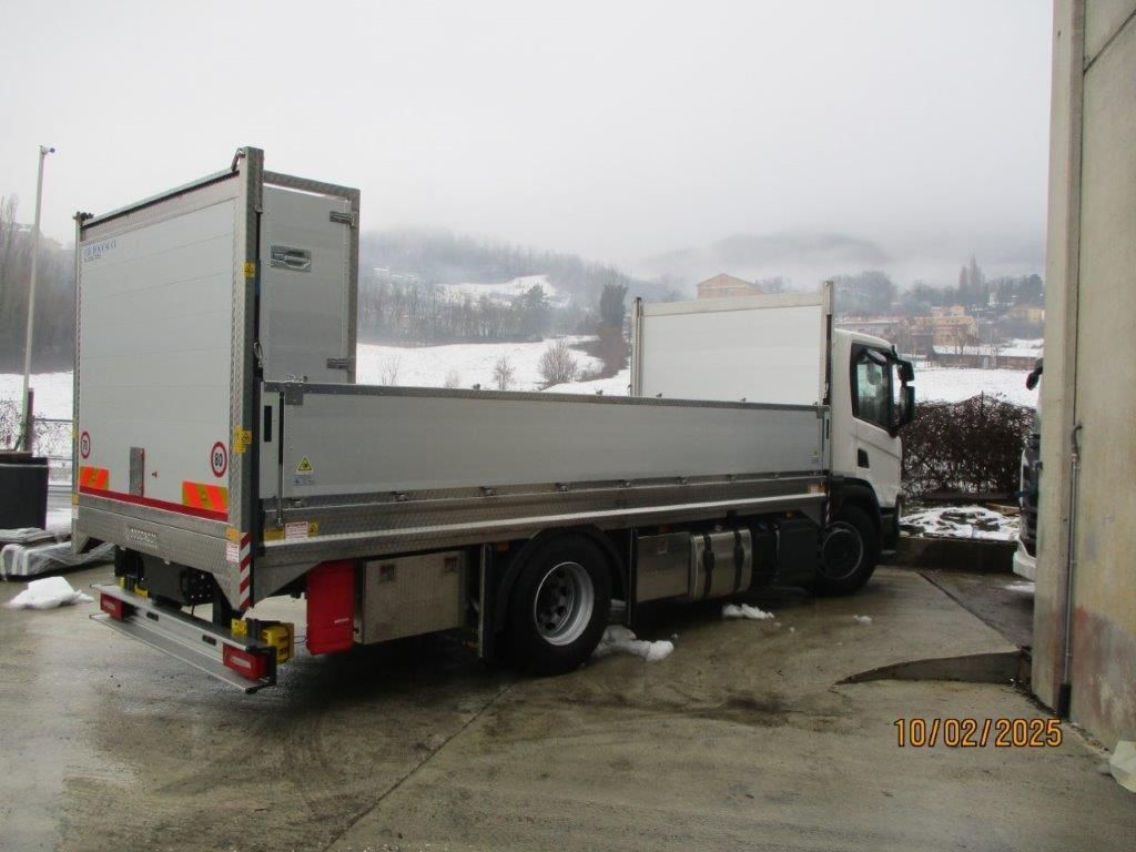 Un camioncino bianco con pianale ribassato è parcheggiato sul cemento, con colline innevate sullo sfondo.