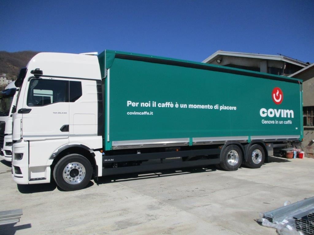 Un camion bianco con un grande rimorchio verde recante il logo del marchio Oovim e scritte in italiano, parcheggiato su un piazzale asfaltato.