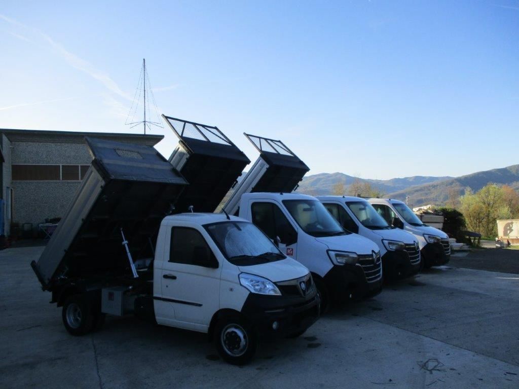 Una fila di autocarri ribaltabili bianchi parcheggiati su un piazzale di cemento sotto un cielo azzurro e limpido, con il cassone del primo camion sollevato.